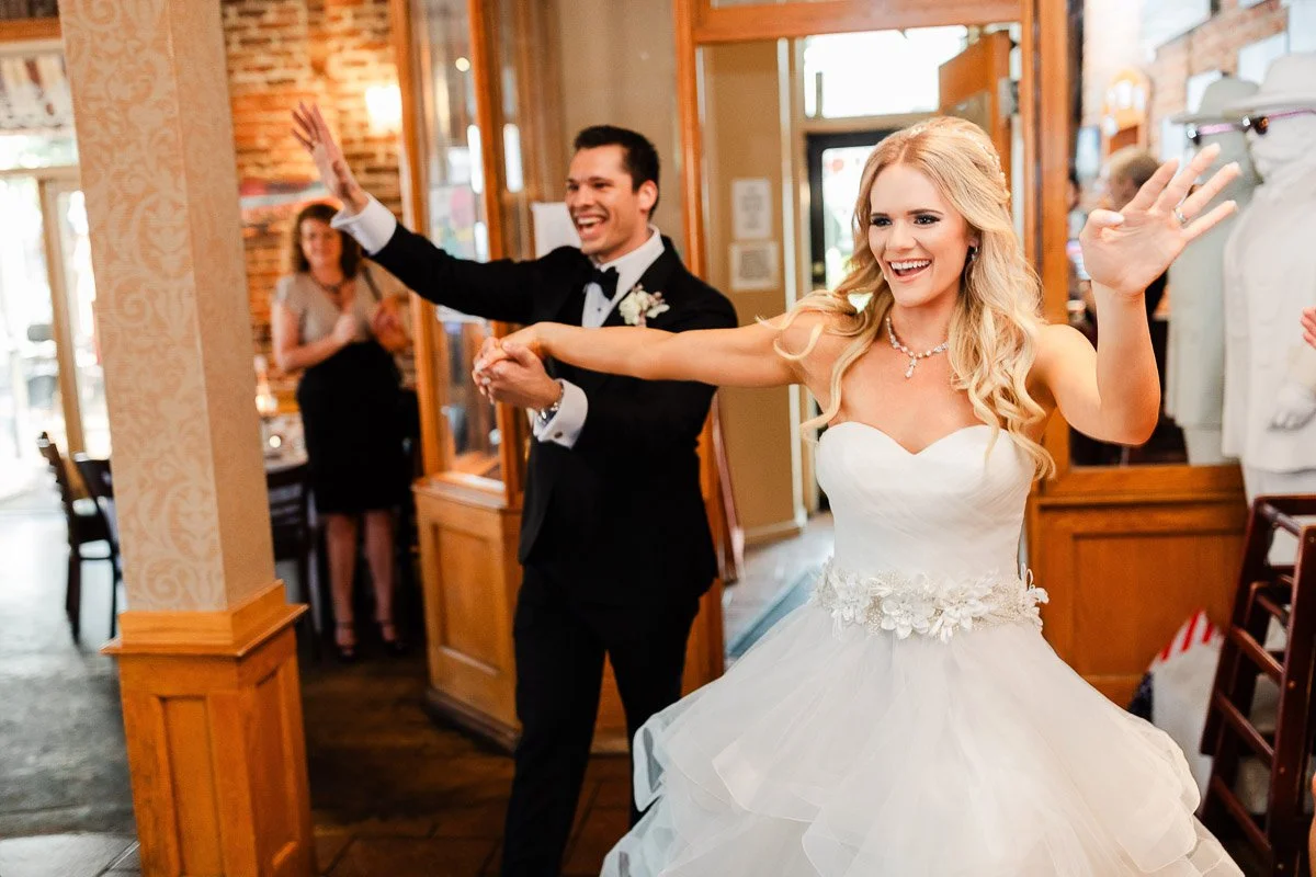 Bride and groom joyfully enter a warmly lit room. The bride wears a white gown, and the groom is in a tuxedo. Guests in the background applause.