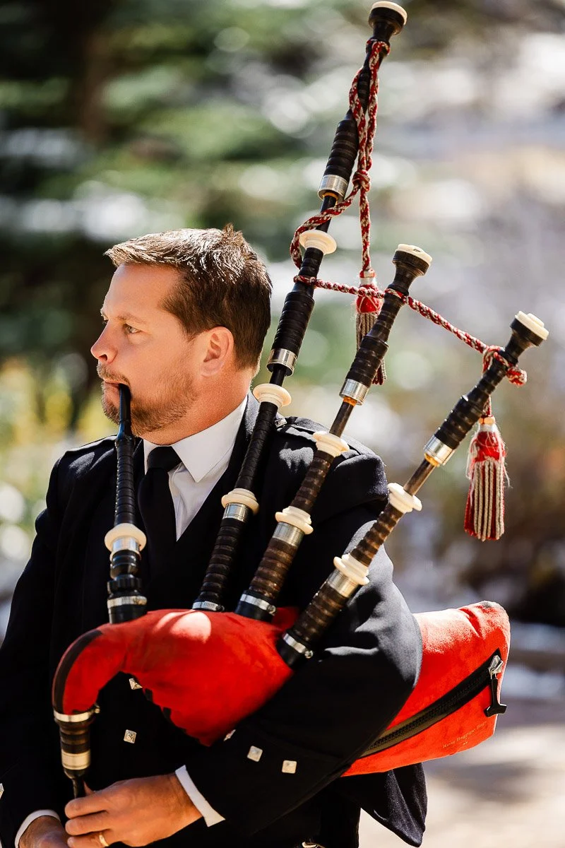A man in formal attire plays a set of red, plaid bagpipes outdoors. Sunlight highlights his concentration, with blurred greenery in the background.