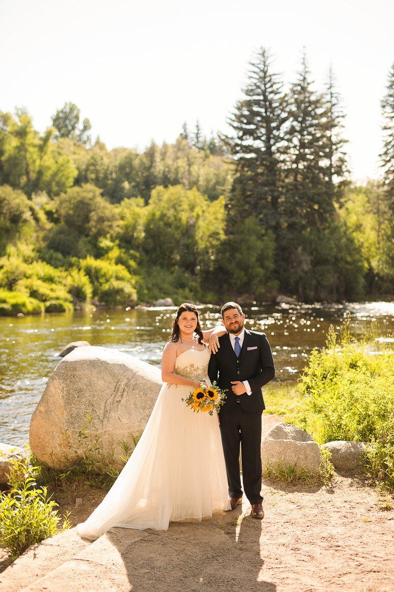 Bride in a flowing white dress and groom in a dark suit standing by a riverbank, holding sunflowers, surrounded by lush greenery and sunlight.