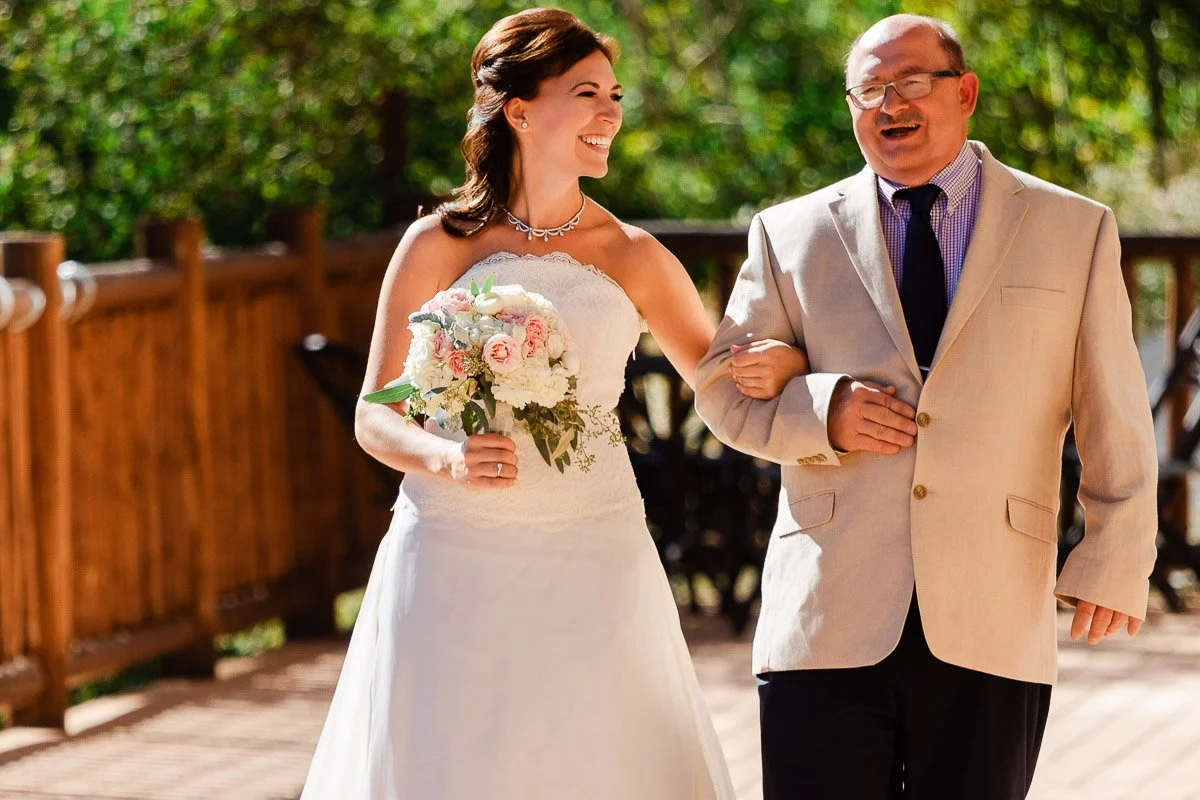 A smiling bride in a white gown holds a bouquet while walking arm-in-arm with an older man in a beige suit on a sunny outdoor wooden deck.