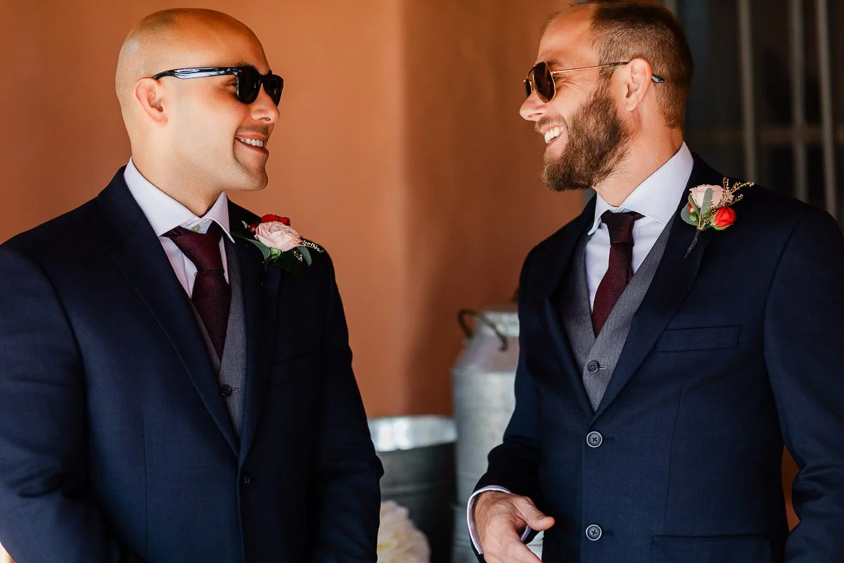 Two men in sunglasses, wearing navy suits with red ties and boutonnières, smile warmly at each other. The background is a soft, peach wall.