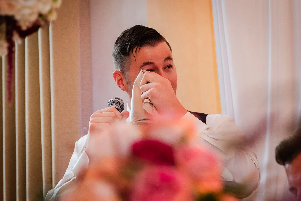 A man in a white dress shirt wipes tears with a napkin while holding a microphone, suggesting an emotional wedding speech. Blurred pink flowers in the foreground.