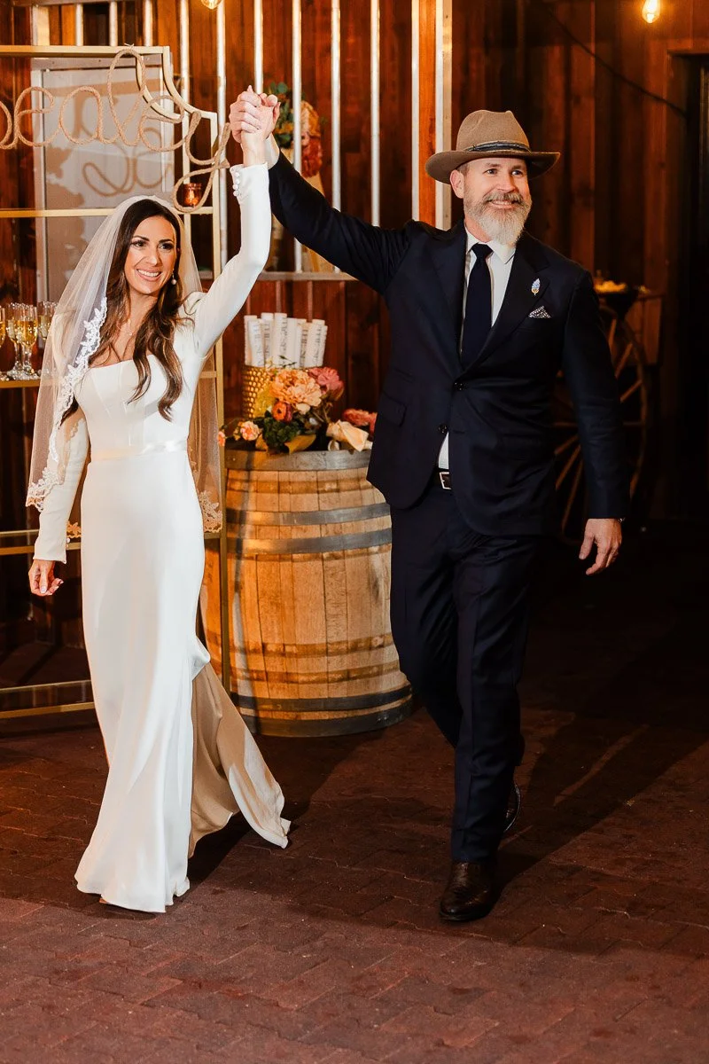 Bride in a white gown and veil, and a groom in a suit and hat, joyously enter a rustic venue with floral decor and warm lighting, holding hands.