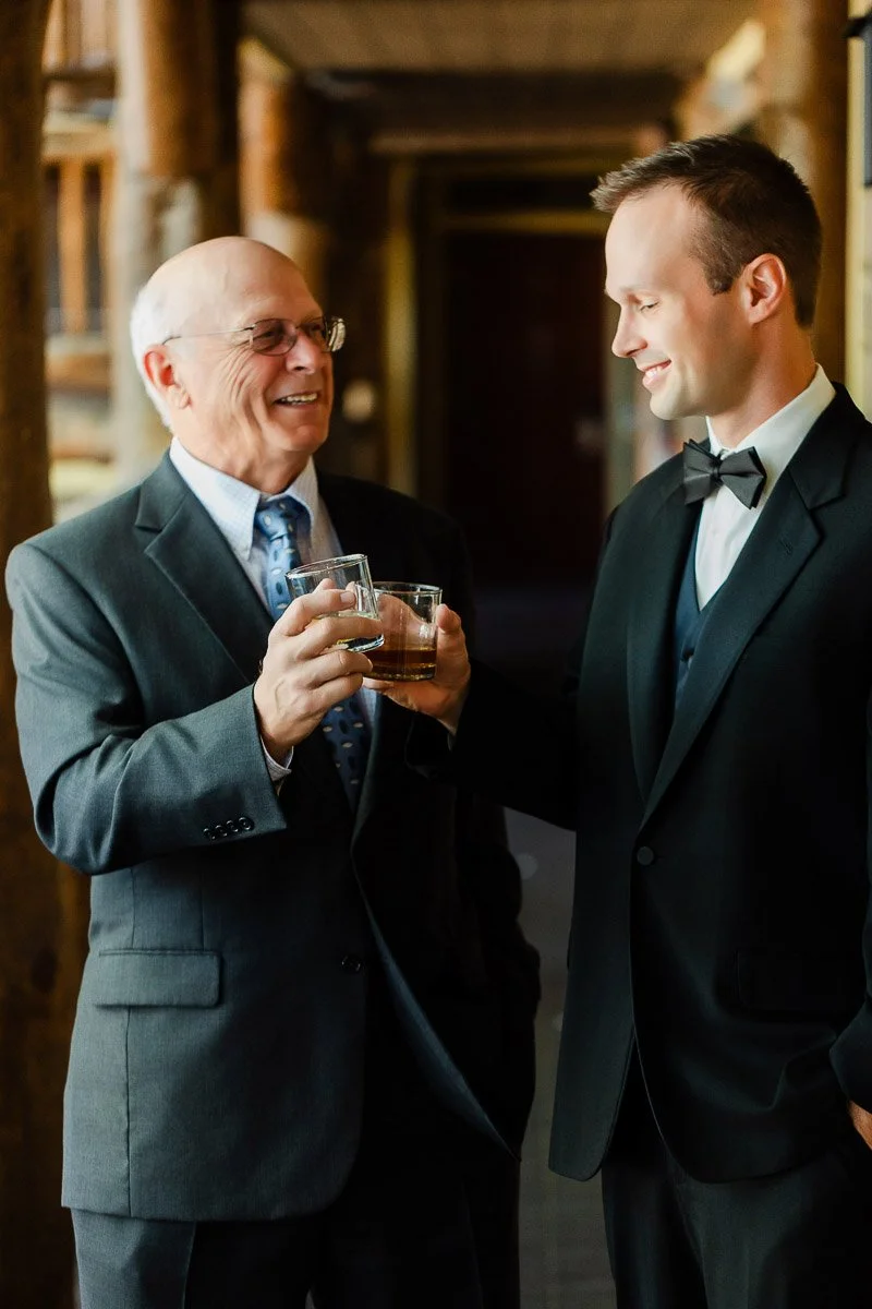 Two men in formal suits clink whiskey glasses, smiling warmly at each other. They stand in a rustic hallway, conveying a celebratory tone.