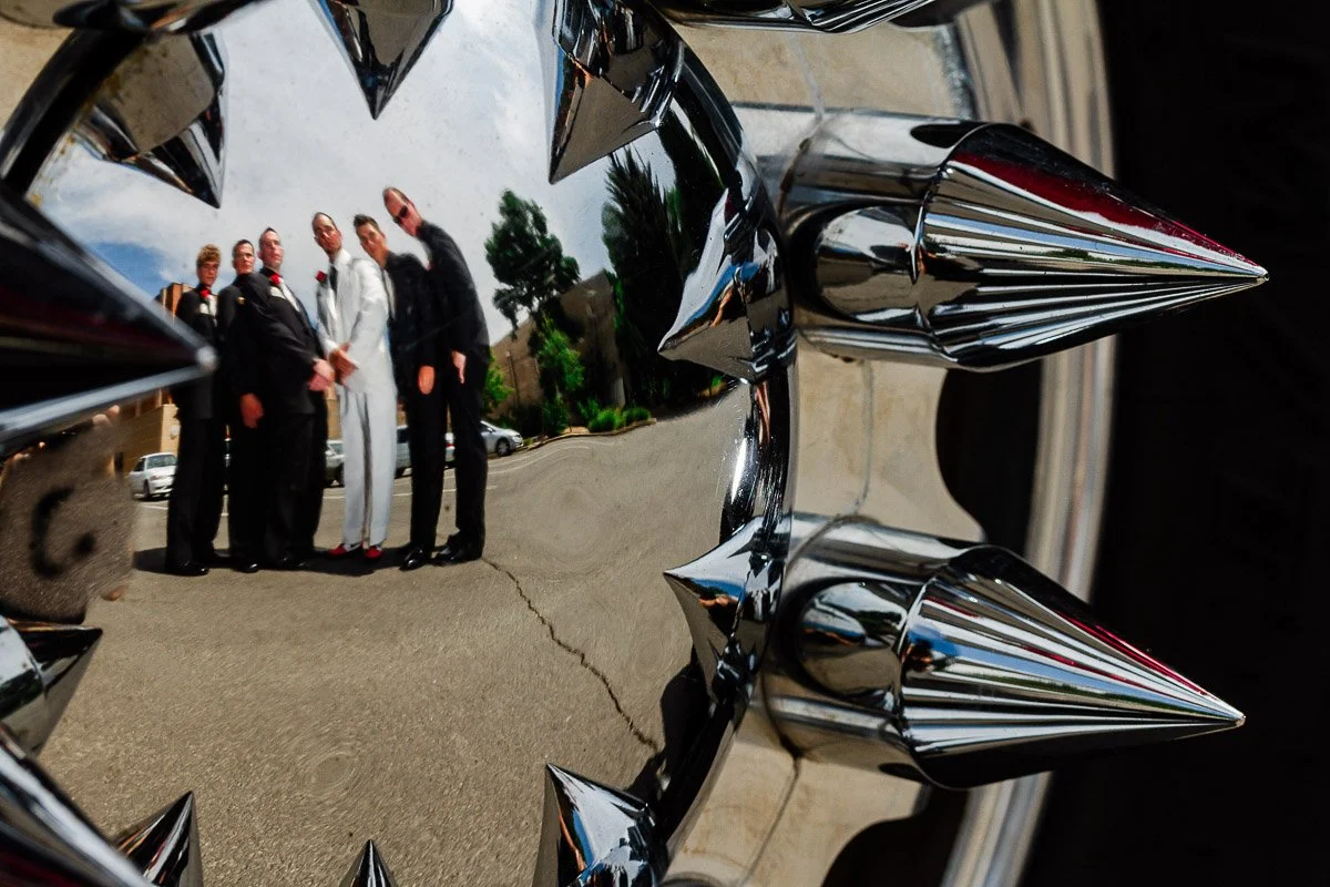Reflection of a group of people in formal attire on a shiny metal surface with pointed edges, against a backdrop of trees and a cloudy sky.