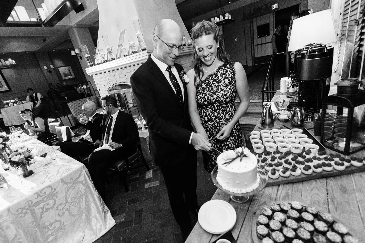A smiling couple in formal attire cuts a cake together at a reception. A decorated table with cupcakes is in the foreground, with guests seated nearby.