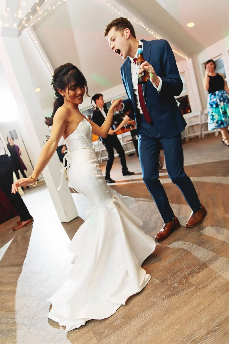 A joyful couple dances at a wedding. The woman, in a flowing white dress, and the man, in a blue suit holding a drink, show energetic happiness.