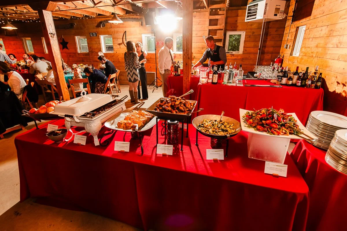 A rustic event hall features a buffet with assorted dishes on red tablecloths. Guests mingle in the background near a bar. Warm, inviting ambiance.