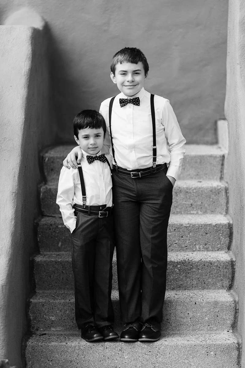 Two young boys in matching outfits stand on steps, smiling. They wear white shirts, black bow ties, suspenders, and dark pants. The tone is cheerful.