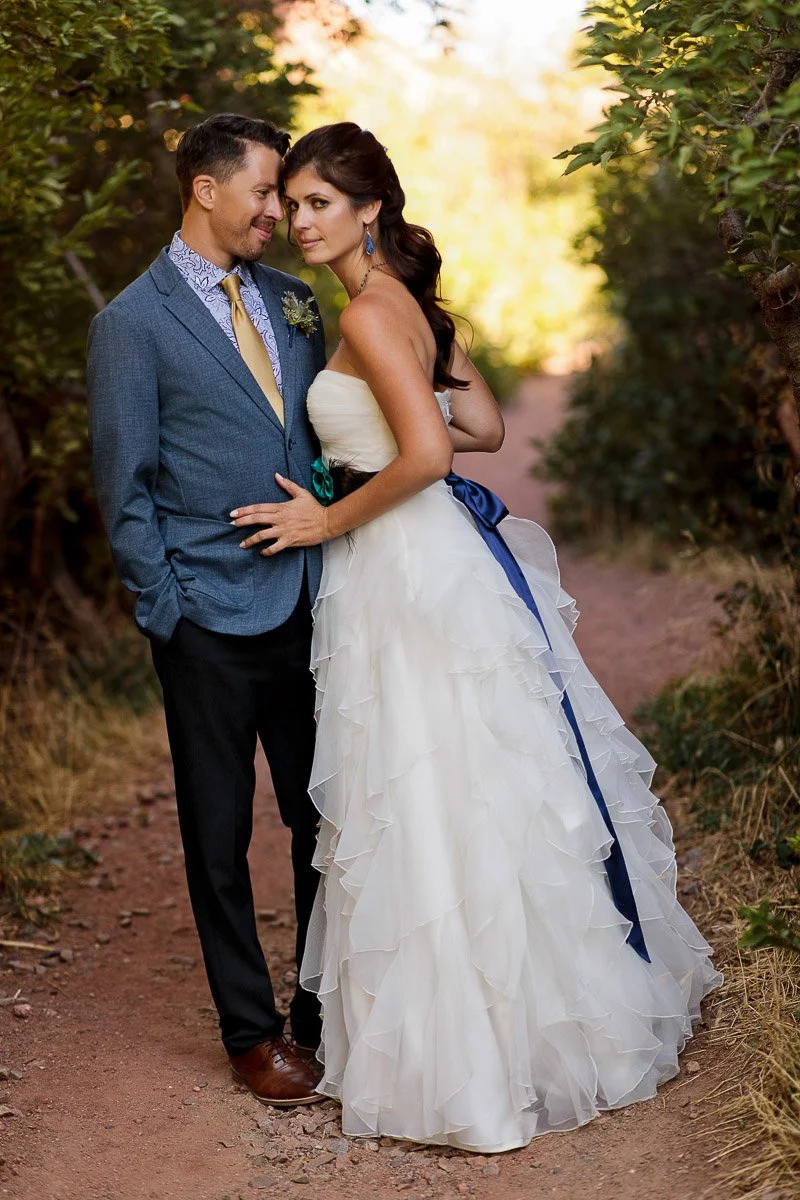 A couple stands on a forest path, warmly embracing. The man wears a blue suit, the woman a ruffled white gown with a navy sash. The scene feels romantic and serene.