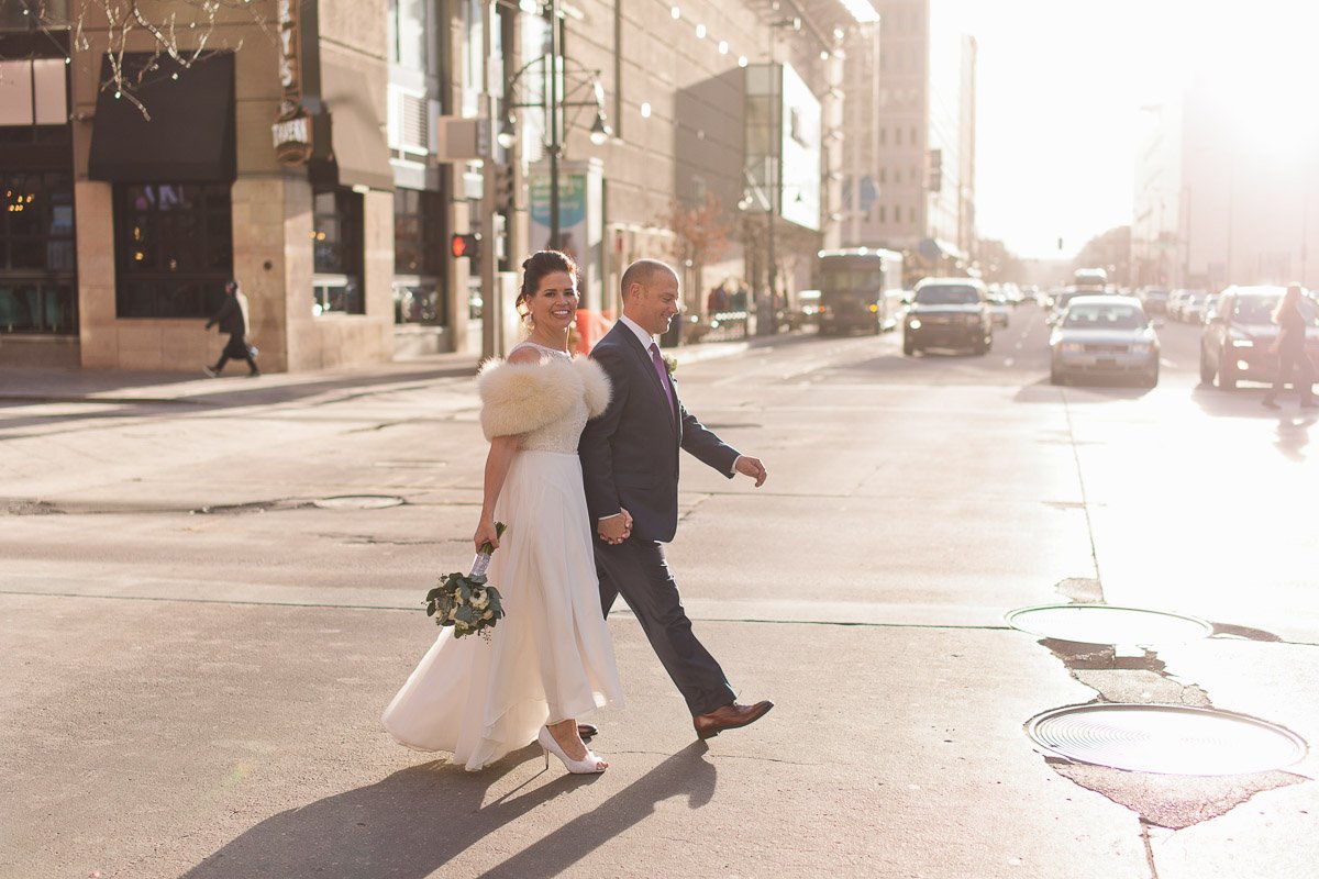 A couple crosses a sunlit city street. The woman wears a white dress and fur shawl, holding flowers. The man is in a suit. The mood is joyful and romantic.