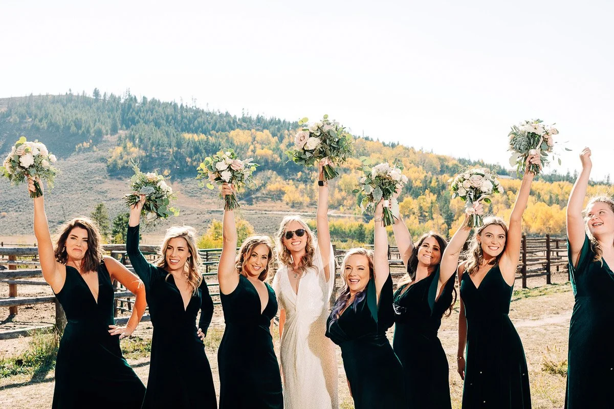 A joyful bride in a white dress and seven bridesmaids in black raise bouquets against a scenic mountainous backdrop with autumn leaves.