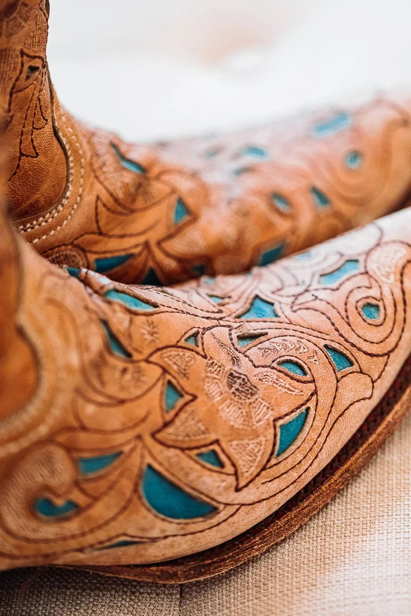 Close-up of tan cowboy boots with intricate floral and scroll embroidery and teal inlays. The boots are resting on a soft, light-colored surface.