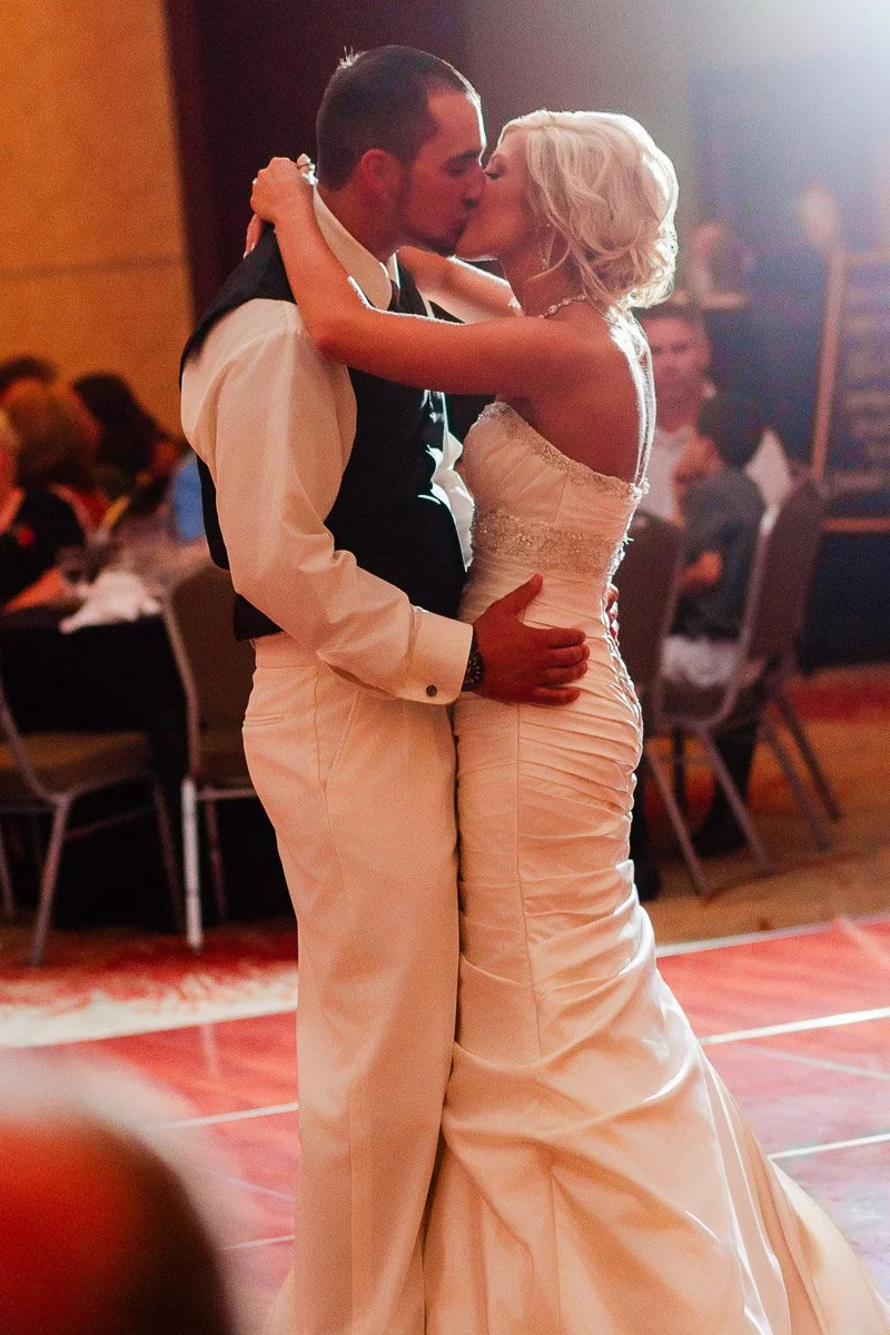 A newlywed couple shares a kiss during their first dance at a wedding reception. The bride wears a white gown, and the groom is in a formal suit. The scene is warm and intimate.