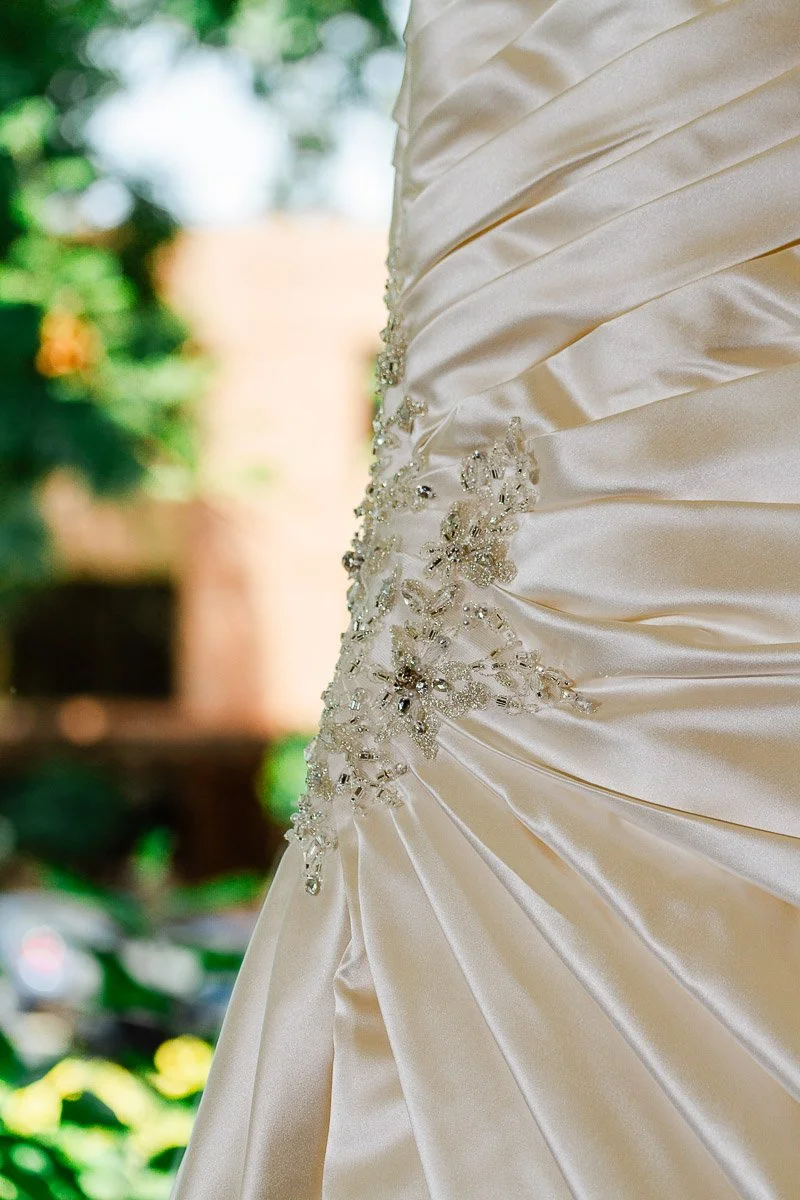 Close-up of an elegant satin wedding dress with intricate beadwork, showcasing its luxurious texture and design. Background hints at a lush, sunny garden.