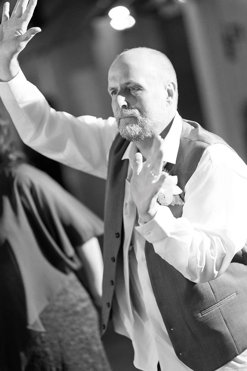 A grayscale image of a bearded man dancing, wearing a vest and shirt, raising his hands joyfully. The setting appears festive and lively.