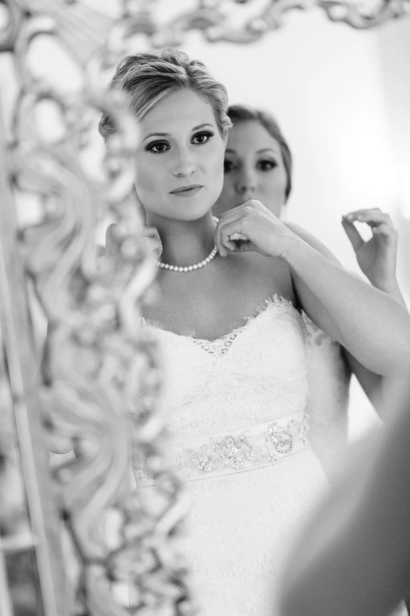 A bride in a lacy white dress and beaded belt gazes thoughtfully in a decorative mirror while someone adjusts her necklace, conveying anticipation and elegance.