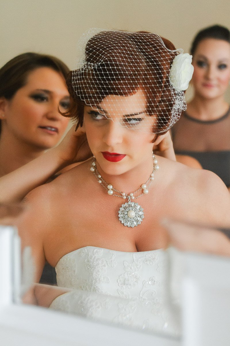 A bride in a white strapless gown stands with two bridesmaids adjusting her veil. She wears a vintage-style necklace, with a calm and focused expression.
