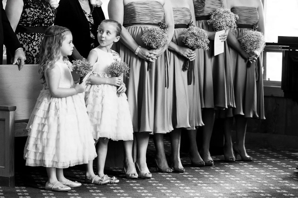Black and white image of a wedding party. Two young flower girls in frilly dresses hold bouquets, standing in front of bridesmaids in mid-length dresses.