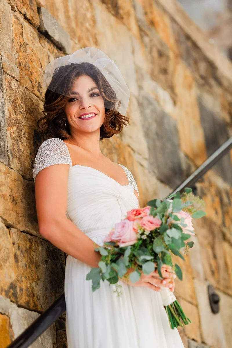 Bride in a white gown with jeweled sleeves, holding a bouquet of pink roses, leans against a stone wall, smiling with a joyful expression.