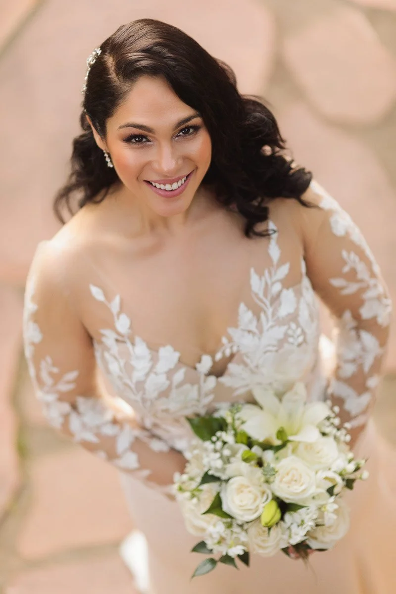 Smiling bride in an embroidered dress holds a bouquet of white roses and lilies. She has wavy brown hair, capturing a joyful and elegant moment.