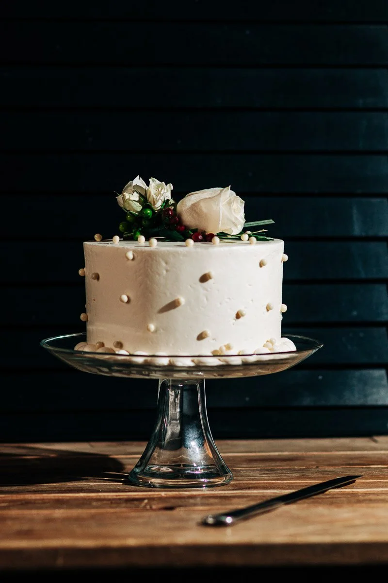 Elegantly decorated white cake with small pearl-like dots, topped with white roses and berries, on a clear stand against a dark background.