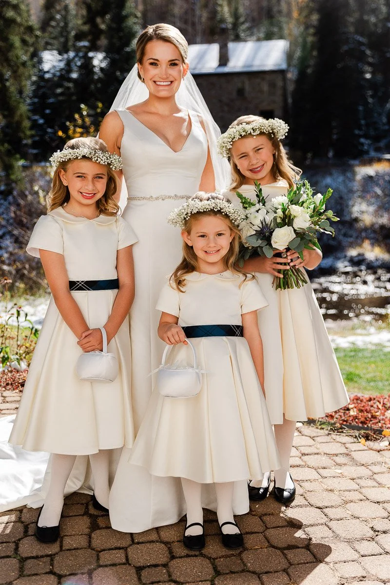 Bride with three flower girls in white dresses pose outdoors. Bride holds bouquet; girls wear floral crowns, smiling on a sunny day near trees and water.