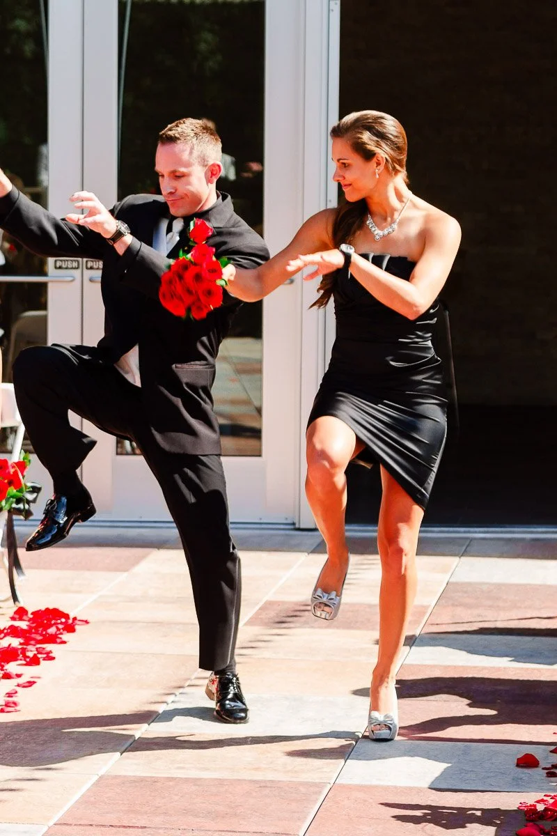A man and woman dance energetically outside. The man wears a black suit and holds roses. The woman wears a black dress. Red petals are scattered on the ground.
