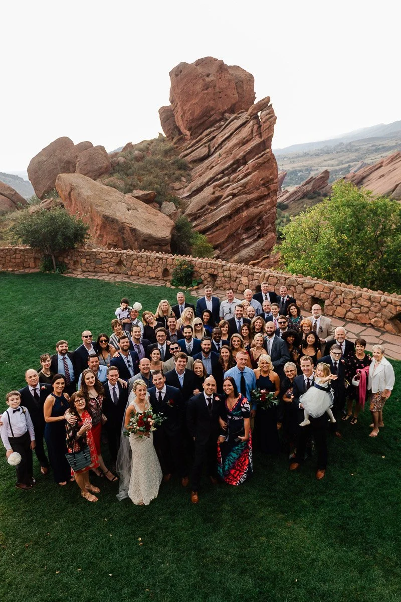 A large wedding group poses on lush green grass with a scenic rocky background. The couple stands smiling at the front, surrounded by joyful, well-dressed guests.