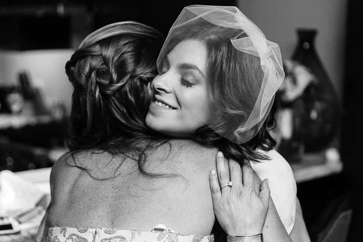 A bride in a veil embraces a woman, smiling with eyes closed, conveying warmth and joy. Black and white photo emphasizes emotion and intimacy.