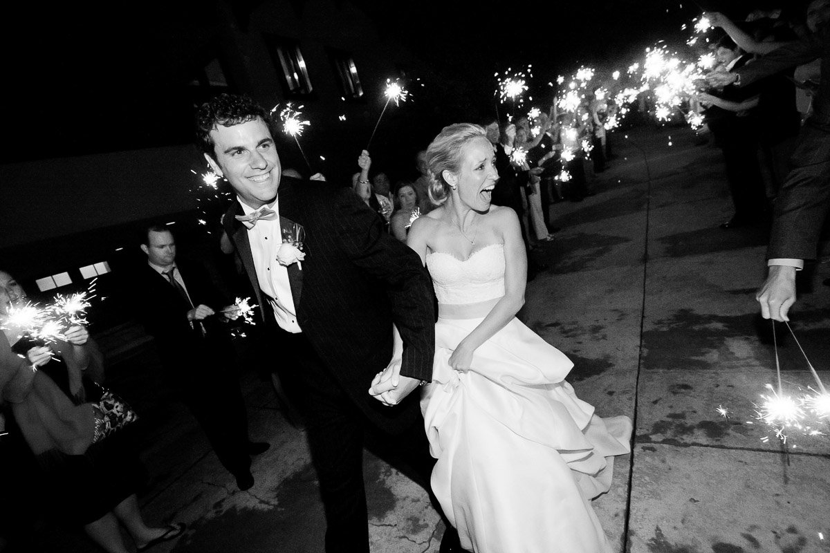 A joyful bride and groom exit their wedding at night, surrounded by guests holding sparklers. The couple is smiling, capturing a moment of celebration.