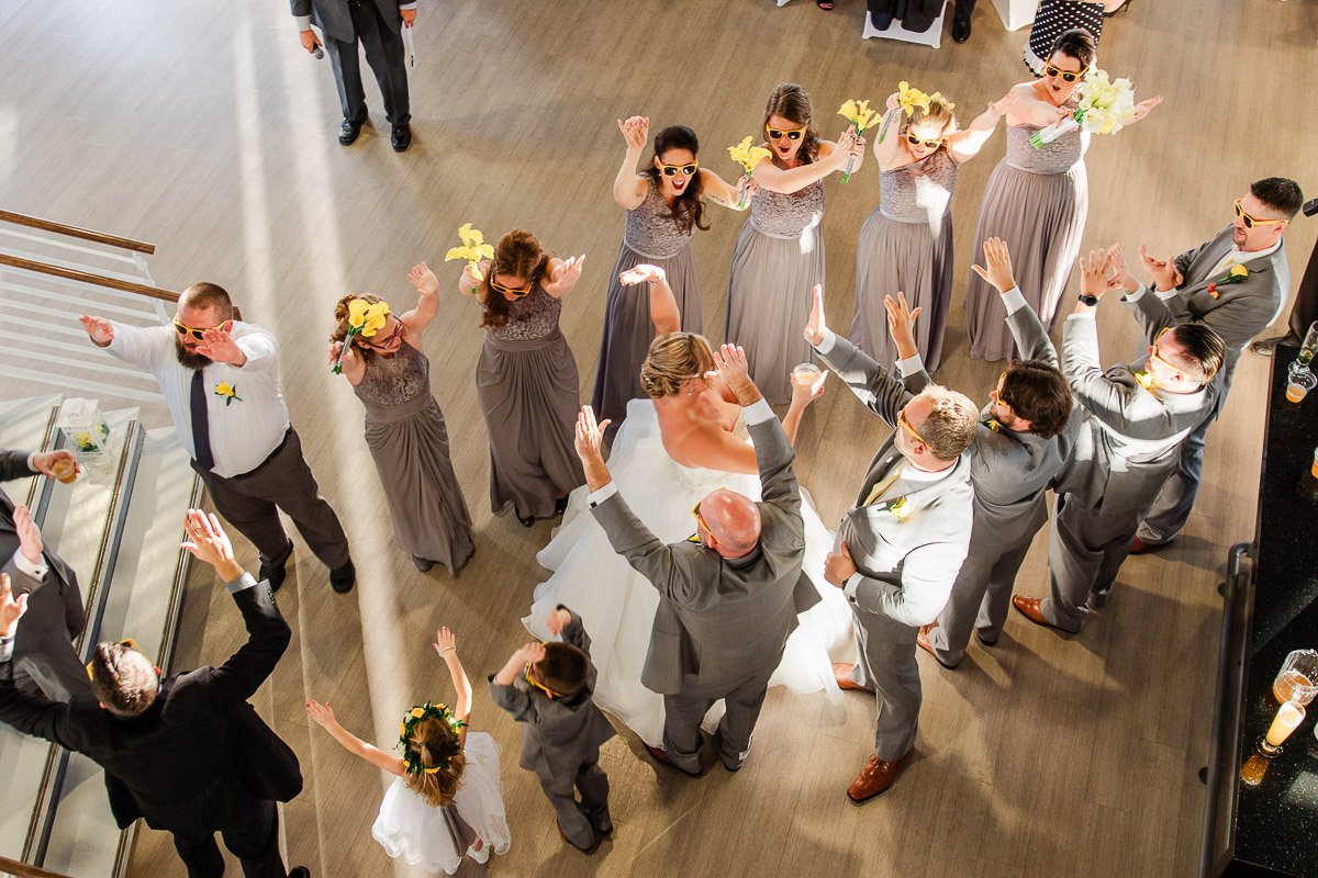 Wedding party in a circle, raising arms joyfully. Bride in white, bridesmaids in gray, wearing sunglasses. Festive atmosphere, wood floor setting.