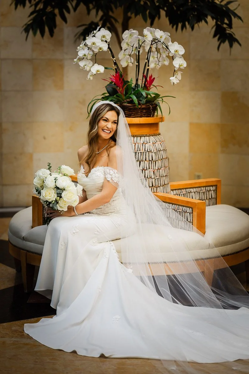 Smiling bride in an elegant white gown sits on a cushioned bench in the lobby of the St Julien Hotel, holding a bouquet of white roses. Orchids in the background add a serene touch.
