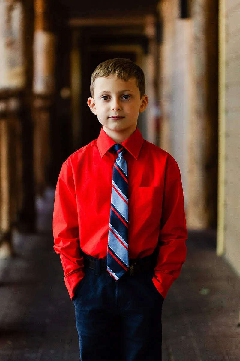 Young boy in a vibrant red shirt and striped tie stands confidently with hands in pockets, set against a warm, softly lit corridor background.