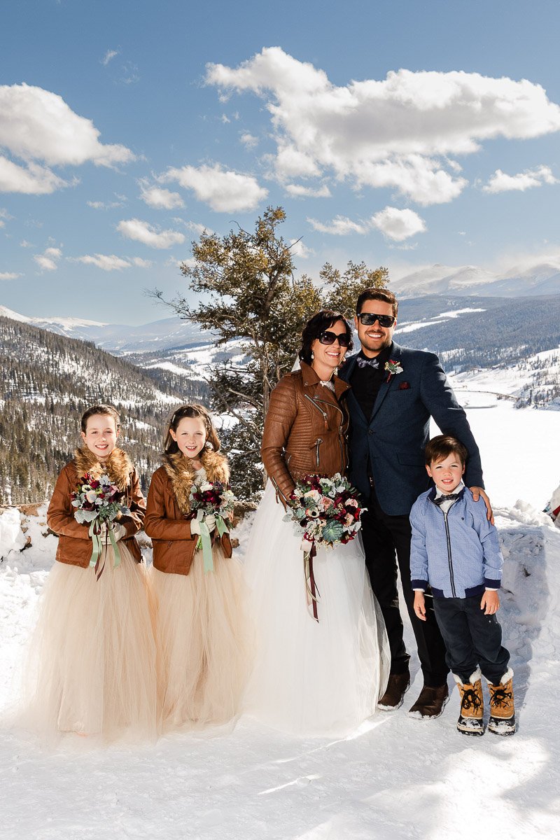 A joyful family stands on a snowy mountain with a stunning view. The parents wear formal clothes; the children in winter attire. Bright, sunny day.