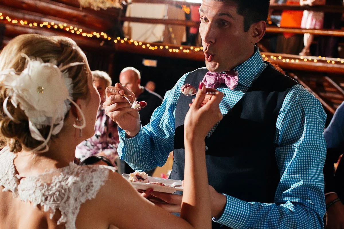 A bride and groom in wedding attire feed each other cake playfully. The setting is festive with warm lighting and a lively atmosphere.