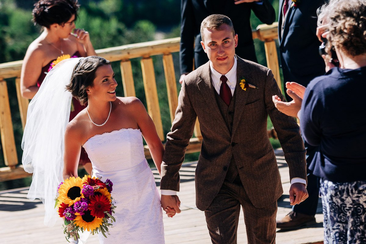 Bride in a white gown and veil, holding sunflowers, smiles while walking hand in hand with a groom in a brown suit. Guests clap in a sunny outdoor setting.
