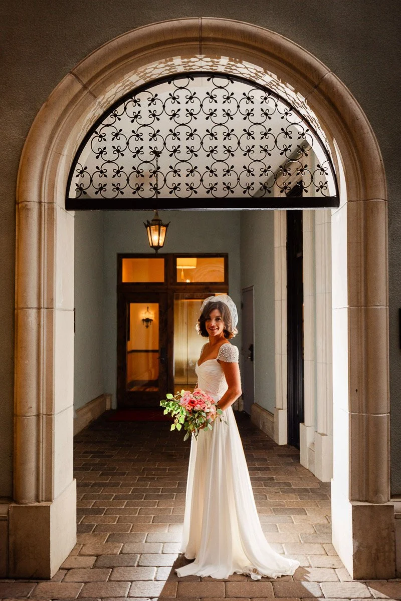 Bride in a sleek white gown stands gracefully in an arched Arrabelle at Vail Square doorway, holding a vibrant bouquet. The scene is elegant and illuminated by soft light.