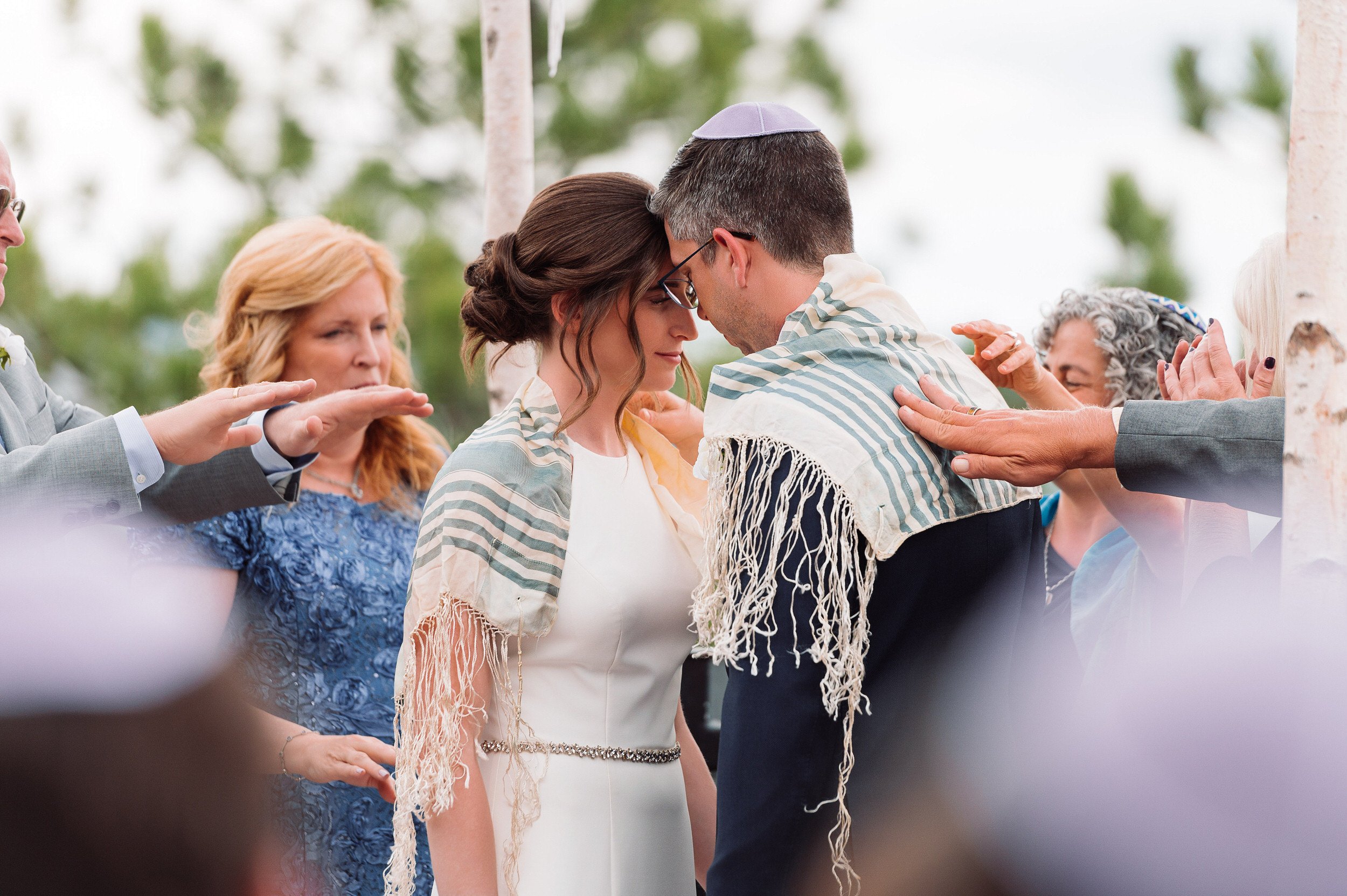 Bride and groom is touched by family under a chuppah during an outdoor wedding ceremony at Mount Vernon Canyon Club in Golden, Colorado.