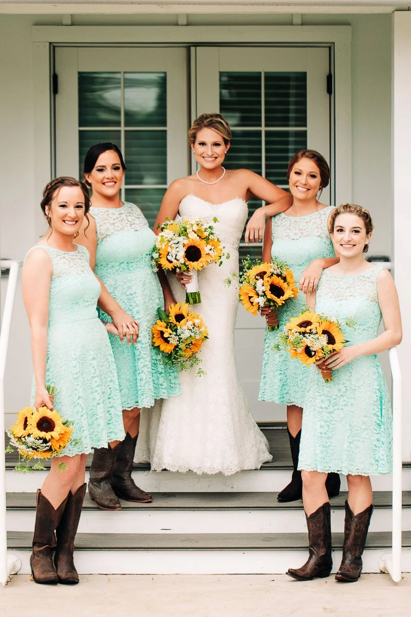 Bride in a white gown and four bridesmaids in mint-green dresses with cowboy boots pose smiling, holding sunflower bouquets on white steps during a Barn at Raccoon Creek wedding in Littleton, Colorado