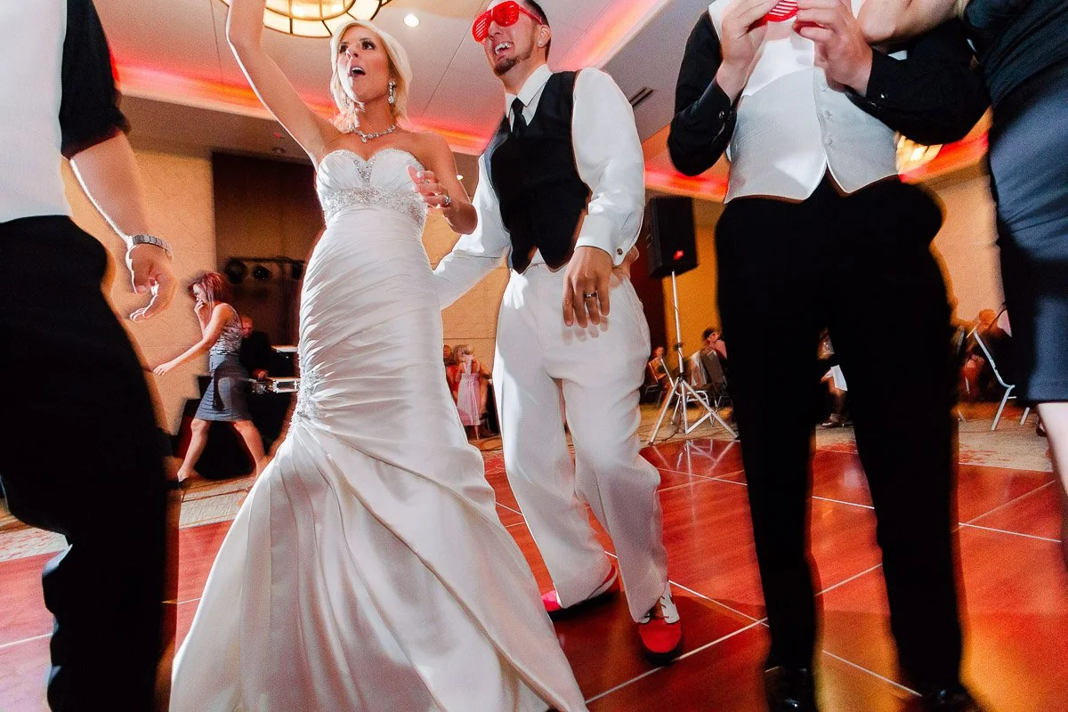 Bride in a strapless gown and groom in a vest and red sunglasses dance joyfully at a wedding reception. Guests around them join the lively celebration.