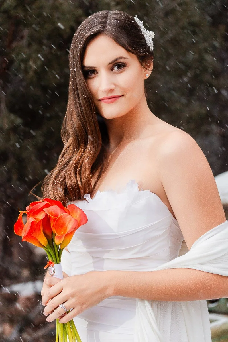 A bride in a strapless white gown holds orange calla lilies. Snow falls gently around her, adding a serene winter feel to the scene.