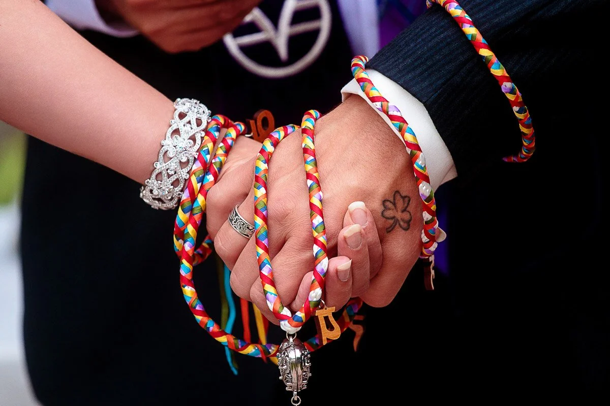Two hands intertwined, wrapped in colorful braided cords, symbolize unity. One hand has a floral tattoo, the other wears a jeweled bracelet.