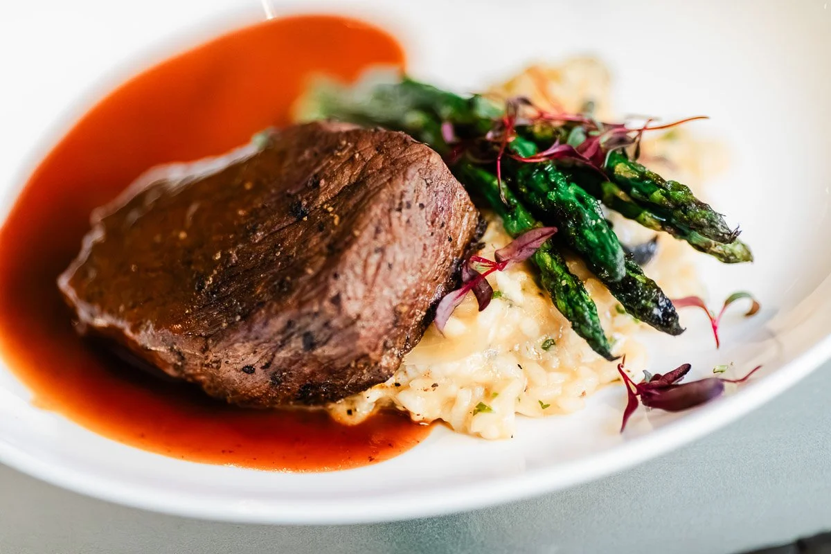 A seared steak rests on creamy risotto, topped with vibrant green asparagus spears. Red wine sauce surrounds the dish, garnished with microgreens.
