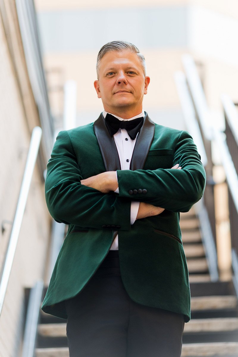 Man in a green velvet tuxedo with crossed arms stands confidently on stairs, conveying elegance and authority. Blurred urban background.