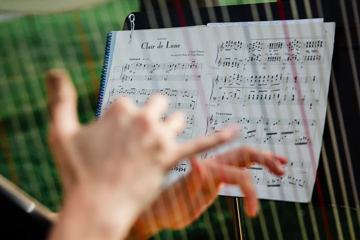 A harpist plays "Clair de Lune," hands gracefully plucking strings, with the open sheet music on a stand and soft outdoor lighting adding elegance.