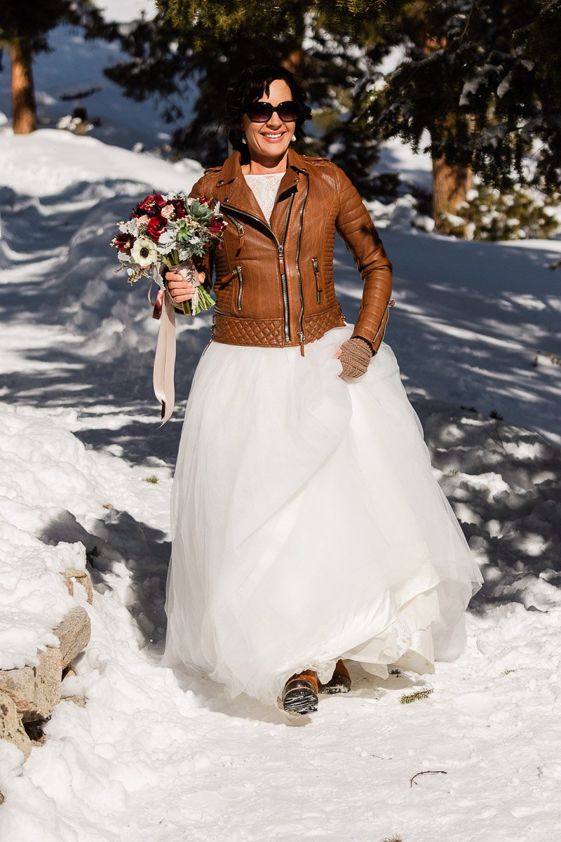 A joyful bride in a leather jacket and white gown walks through snowy woods, holding a vibrant bouquet. The scene is bright and wintry.