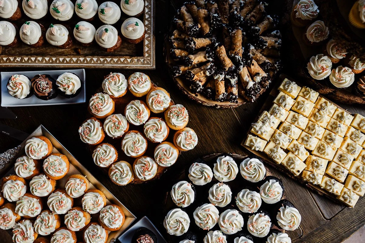 Assorted desserts on a dark table, featuring cupcakes with frosting, sprinkled cannoli, and neatly arranged cheesecake squares, creating a festive display.