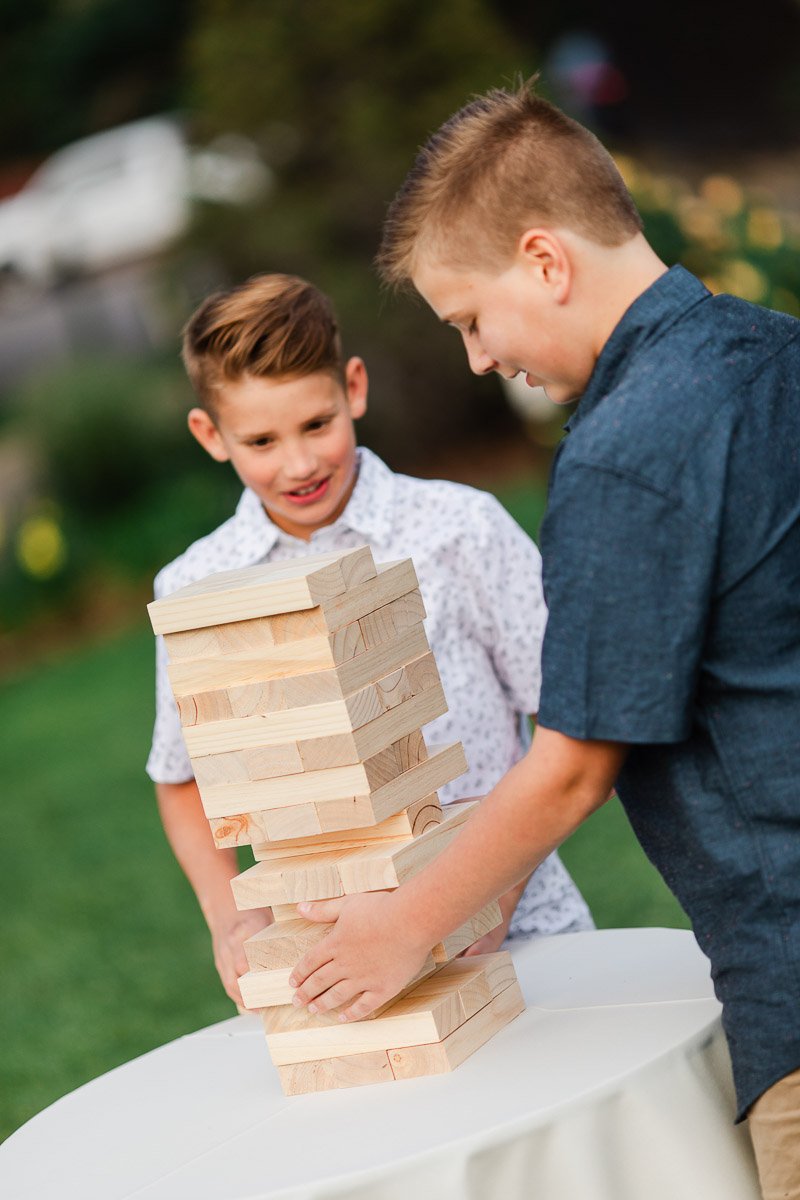 Two boys play an outdoor game with large wooden blocks on a table. One carefully pulls a block while the other watches, both focused and smiling.