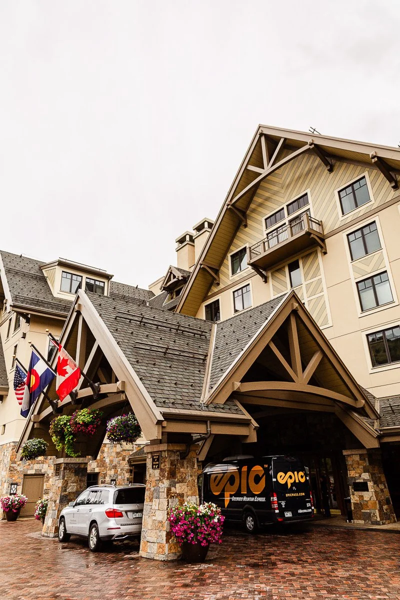 Four Seasons Resort lodge entrance in Vail Colorado with peaked roofs, a U.S., Colorado, and Canadian flag, adorned with flower baskets. A white car and a van are parked.